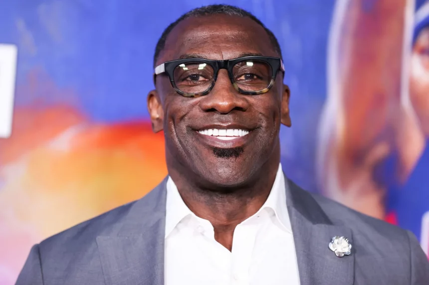Shannon Sharpe mit schwarzer Brille, grauem Jackett und weißem Hemd steht vor einem bunten, unscharfen Hintergrund. Er hat kurze Haare, einen Spitzbart und eine weiße Blumenbrosche – eine Reminiszenz an Shannon Sharpes klassischen Stil. Diese Beschreibung wurde mit der FootballR KI automatisch generiert.