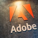 NFL und Adobe - Das Bild zeigt das Adobe Logo: ein stilisiertes rotes „A“, das aus geometrischen Formen über dem Wort „Adobe“ in fetten weißen Buchstaben gebildet wird, angezeigt auf einem strukturierten, dunkelgrauen Hintergrund unter warmer Beleuchtung, was die KI-gestützte Partnerschaft von Adobe mit der NFL hervorhebt. Diese Beschreibung wurde mit der FootballR KI automatisch generiert.