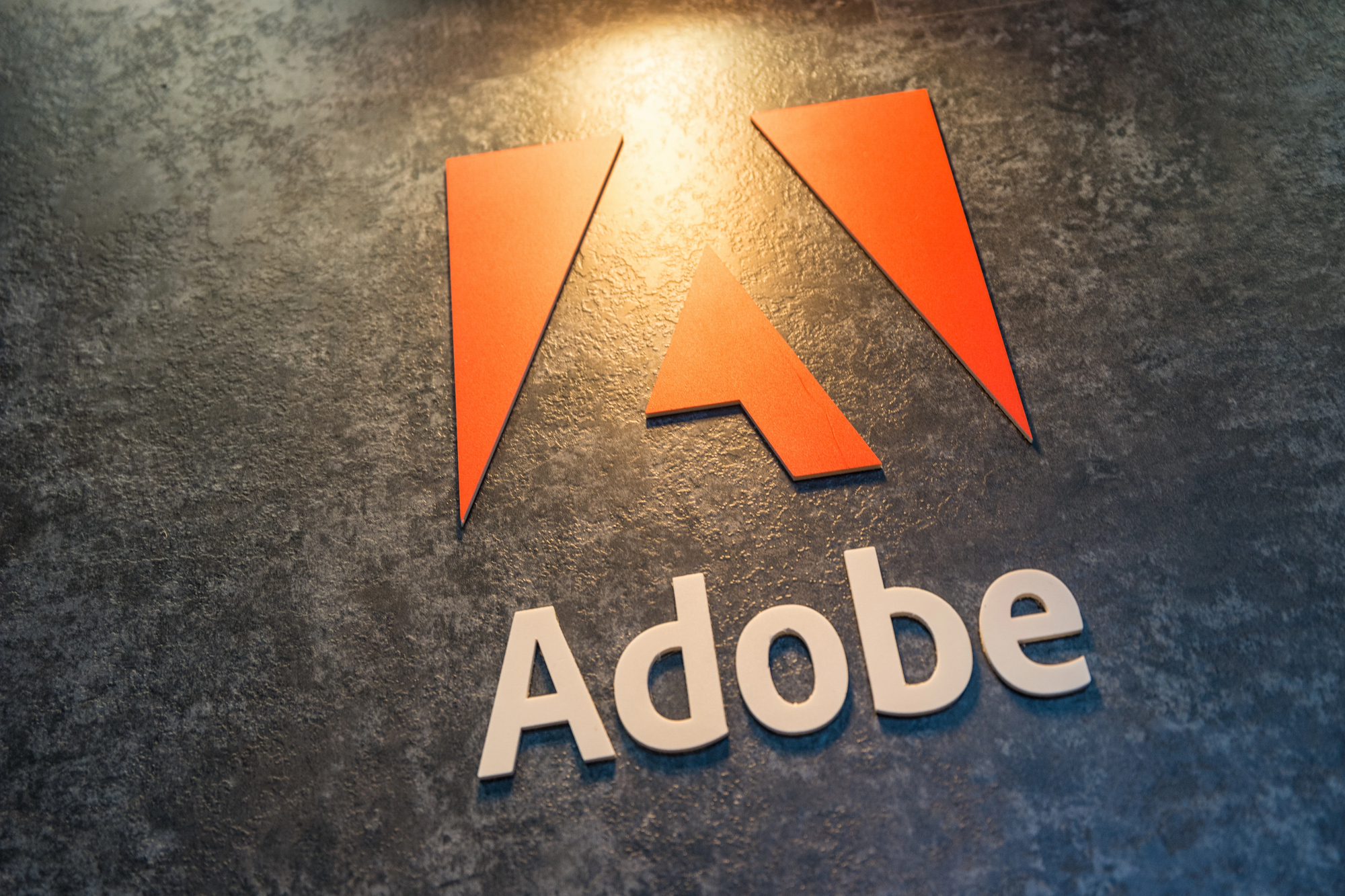 NFL und Adobe - Das Bild zeigt das Adobe Logo: ein stilisiertes rotes „A“, das aus geometrischen Formen über dem Wort „Adobe“ in fetten weißen Buchstaben gebildet wird, angezeigt auf einem strukturierten, dunkelgrauen Hintergrund unter warmer Beleuchtung, was die KI-gestützte Partnerschaft von Adobe mit der NFL hervorhebt. Diese Beschreibung wurde mit der FootballR KI automatisch generiert.
