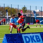 Zwei Sportler treten in einem NFL-FLAG-Footballspiel auf einem leuchtend grünen Feld an. Einer im blauen Trikot weicht einem anderen Spieler im roten Trikot aus. Bunte Banner und Gebäude weisen auf Deutschland hin, während im Vordergrund ein „DKB“-Schild sticht. Diese Beschreibung wurde mit der FootballR KI automatisch generiert.