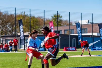 Zwei Sportler treten in einem NFL-FLAG-Footballspiel auf einem leuchtend grünen Feld an. Einer im blauen Trikot weicht einem anderen Spieler im roten Trikot aus. Bunte Banner und Gebäude weisen auf Deutschland hin, während im Vordergrund ein „DKB“-Schild sticht. Diese Beschreibung wurde mit der FootballR KI automatisch generiert.