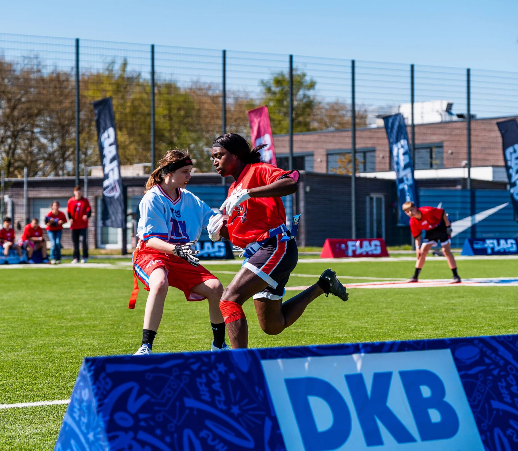 Zwei Sportler treten in einem NFL-FLAG-Footballspiel auf einem leuchtend grünen Feld an. Einer im blauen Trikot weicht einem anderen Spieler im roten Trikot aus. Bunte Banner und Gebäude weisen auf Deutschland hin, während im Vordergrund ein „DKB“-Schild sticht. Diese Beschreibung wurde mit der FootballR KI automatisch generiert.