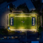 Eine nächtliche Luftaufnahme zeigt ein Footballfeld, hell erleuchtet von Flutlicht. Das Patriots-Logo befindet sich in der Mitte und wird an beiden Enden gespiegelt. Schwach beleuchtete Gebäude umgeben dieses Flag-Football-Stadion und lassen die New England Patriots mit ihrer lebendigen Beleuchtung im Mittelpunkt erstrahlen. Diese Beschreibung wurde mit der FootballR KI automatisch generiert.