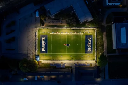 Eine nächtliche Luftaufnahme zeigt ein Footballfeld, hell erleuchtet von Flutlicht. Das Patriots-Logo befindet sich in der Mitte und wird an beiden Enden gespiegelt. Schwach beleuchtete Gebäude umgeben dieses Flag-Football-Stadion und lassen die New England Patriots mit ihrer lebendigen Beleuchtung im Mittelpunkt erstrahlen. Diese Beschreibung wurde mit der FootballR KI automatisch generiert.