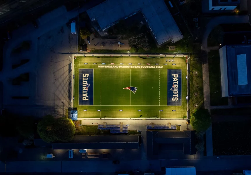 Eine nächtliche Luftaufnahme zeigt ein Footballfeld, hell erleuchtet von Flutlicht. Das Patriots-Logo befindet sich in der Mitte und wird an beiden Enden gespiegelt. Schwach beleuchtete Gebäude umgeben dieses Flag-Football-Stadion und lassen die New England Patriots mit ihrer lebendigen Beleuchtung im Mittelpunkt erstrahlen. Diese Beschreibung wurde mit der FootballR KI automatisch generiert.