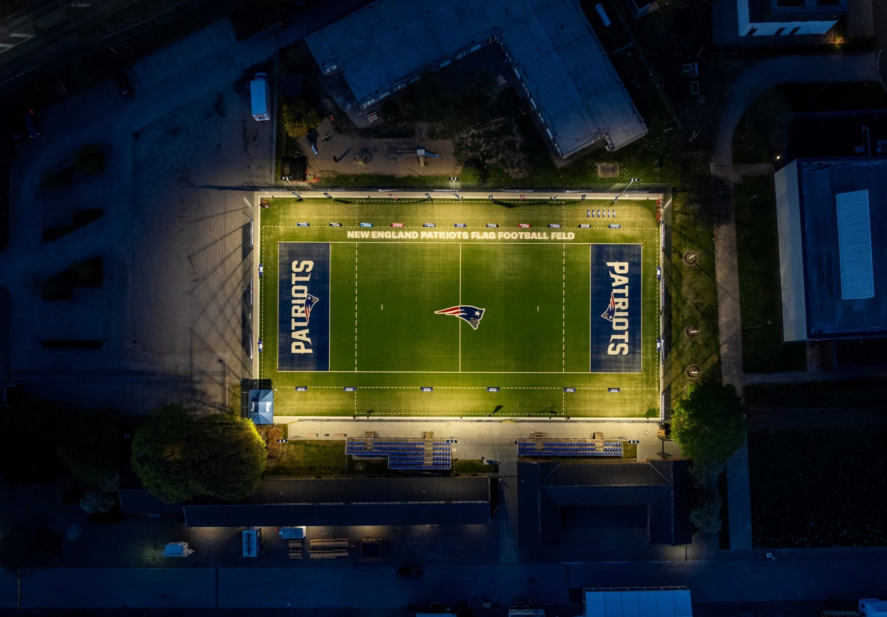 Eine nächtliche Luftaufnahme zeigt ein Footballfeld, hell erleuchtet von Flutlicht. Das Patriots-Logo befindet sich in der Mitte und wird an beiden Enden gespiegelt. Schwach beleuchtete Gebäude umgeben dieses Flag-Football-Stadion und lassen die New England Patriots mit ihrer lebendigen Beleuchtung im Mittelpunkt erstrahlen. Diese Beschreibung wurde mit der FootballR KI automatisch generiert.