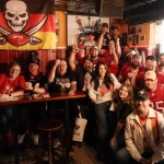Eine lebhafte Gruppe von Tampa Bay Buccaneers-Fans in Rot, Schwarz und Weiß posiert in einem gemütlichen Pub. Sie lächeln, jubeln und halten ihre Teamkleidung hoch, während hinter ihnen eine Totenkopf-Football-Flagge hängt. Holztische, gerahmte Fotos und gedämpftes Licht schaffen die passende Atmosphäre. Diese Beschreibung wurde mit der FootballR KI automatisch generiert.
