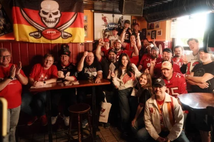 Eine lebhafte Gruppe von Tampa Bay Buccaneers-Fans in Rot, Schwarz und Weiß posiert in einem gemütlichen Pub. Sie lächeln, jubeln und halten ihre Teamkleidung hoch, während hinter ihnen eine Totenkopf-Football-Flagge hängt. Holztische, gerahmte Fotos und gedämpftes Licht schaffen die passende Atmosphäre. Diese Beschreibung wurde mit der FootballR KI automatisch generiert.