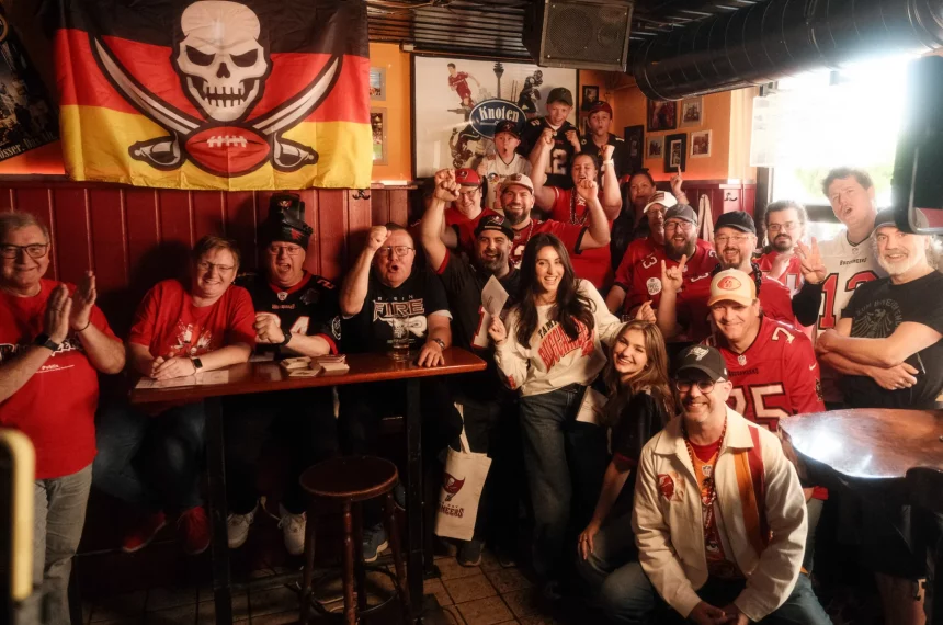 Eine lebhafte Gruppe von Tampa Bay Buccaneers-Fans in Rot, Schwarz und Weiß posiert in einem gemütlichen Pub. Sie lächeln, jubeln und halten ihre Teamkleidung hoch, während hinter ihnen eine Totenkopf-Football-Flagge hängt. Holztische, gerahmte Fotos und gedämpftes Licht schaffen die passende Atmosphäre. Diese Beschreibung wurde mit der FootballR KI automatisch generiert.