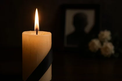 Im Vordergrund steht eine brennende Kerze mit schwarzem Band, die Trauer symbolisiert. Im unscharfen Hintergrund sind die gerahmte Silhouette von Don Hasselbeck und weiße Blumen zu sehen, die vor der dunklen Kulisse eine düstere Atmosphäre schaffen. Diese Beschreibung wurde mit der FootballR KI automatisch generiert.