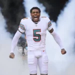 Jalen Ramsey, in der weißen Uniform der Miami Dolphins mit der Nummer 5, steht inmitten von aufsteigendem Rauch, die Fäuste geballt und schreiend, während Fotografen im Hintergrund die dramatische Vorstellung des Spielers einfangen. Diese Beschreibung wurde mit der FootballR KI automatisch generiert.
