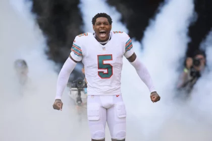 Jalen Ramsey, in der weißen Uniform der Miami Dolphins mit der Nummer 5, steht inmitten von aufsteigendem Rauch, die Fäuste geballt und schreiend, während Fotografen im Hintergrund die dramatische Vorstellung des Spielers einfangen. Diese Beschreibung wurde mit der FootballR KI automatisch generiert.