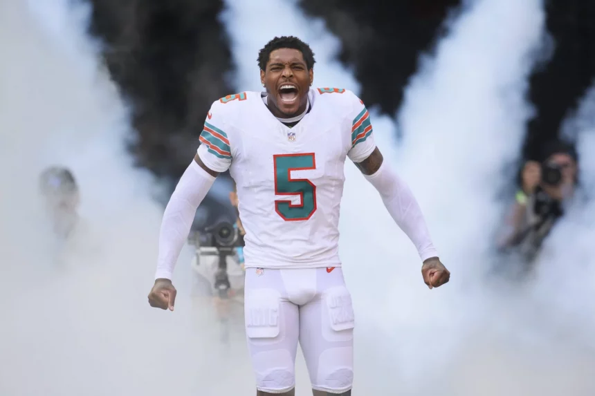 Jalen Ramsey, in der weißen Uniform der Miami Dolphins mit der Nummer 5, steht inmitten von aufsteigendem Rauch, die Fäuste geballt und schreiend, während Fotografen im Hintergrund die dramatische Vorstellung des Spielers einfangen. Diese Beschreibung wurde mit der FootballR KI automatisch generiert.