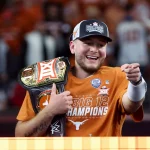 Ein Mann in einem orangefarbenen „Big 12 Champions“-Shirt und einer Baseballkappe lächelt und zeigt in die Kamera. Er verkörpert die Begeisterung einer siegreichen Saison. Mit einem Meisterschaftsgürtel über der Schulter strahlt er die Energie von Quinn Ewers aus. Im Hintergrund summt eine verschwommene Menschenmenge in passenden Farben. Diese Beschreibung wurde mit der FootballR KI automatisch generiert.