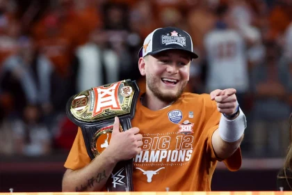 Ein Mann in einem orangefarbenen „Big 12 Champions“-Shirt und einer Baseballkappe lächelt und zeigt in die Kamera. Er verkörpert die Begeisterung einer siegreichen Saison. Mit einem Meisterschaftsgürtel über der Schulter strahlt er die Energie von Quinn Ewers aus. Im Hintergrund summt eine verschwommene Menschenmenge in passenden Farben. Diese Beschreibung wurde mit der FootballR KI automatisch generiert.