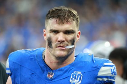Ein Footballspieler, John Cominsky, trägt ein blaues Trikot mit weißen und schwarzen Markierungen und blickt in die Kamera. Sein Gesicht ist mit verschmierter schwarzer Augenfarbe geschmückt. Das Trikot zeigt ein NFL-Logo und einen „90 Seasons“-Aufnäher. Er strahlt Konzentration und Intensität aus, sein Mundschutz ist teilweise sichtbar. Diese Beschreibung wurde mit der FootballR KI automatisch generiert.