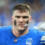 Ein Footballspieler, John Cominsky, trägt ein blaues Trikot mit weißen und schwarzen Markierungen und blickt in die Kamera. Sein Gesicht ist mit verschmierter schwarzer Augenfarbe geschmückt. Das Trikot zeigt ein NFL-Logo und einen „90 Seasons“-Aufnäher. Er strahlt Konzentration und Intensität aus, sein Mundschutz ist teilweise sichtbar. Diese Beschreibung wurde mit der FootballR KI automatisch generiert.