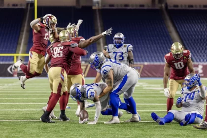 Ein Footballspieler in Blau und Weiß wird angegriffen und verliert den Ball, während vier Spieler in Rot und Gold verteidigen. Ein rot gekleideter Spieler springt mit erhobenen Armen, ein anderer greift nach dem Ball – Nacua schlägt Fan und sorgt für Skandal in den leeren Stadionsitzen. Diese Beschreibung wurde mit der FootballR KI automatisch generiert.
