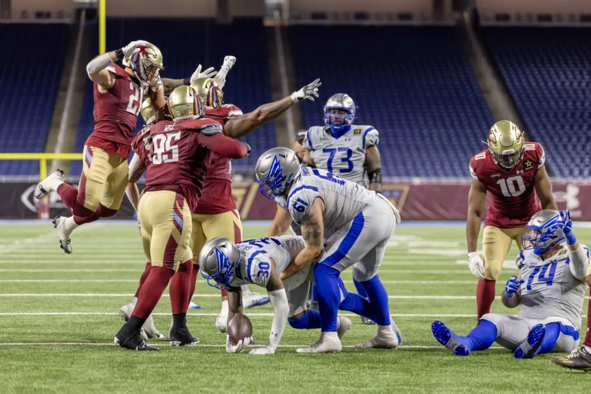 Ein Footballspieler in Blau und Weiß wird angegriffen und verliert den Ball, während vier Spieler in Rot und Gold verteidigen. Ein rot gekleideter Spieler springt mit erhobenen Armen, ein anderer greift nach dem Ball – Nacua schlägt Fan und sorgt für Skandal in den leeren Stadionsitzen. Diese Beschreibung wurde mit der FootballR KI automatisch generiert.