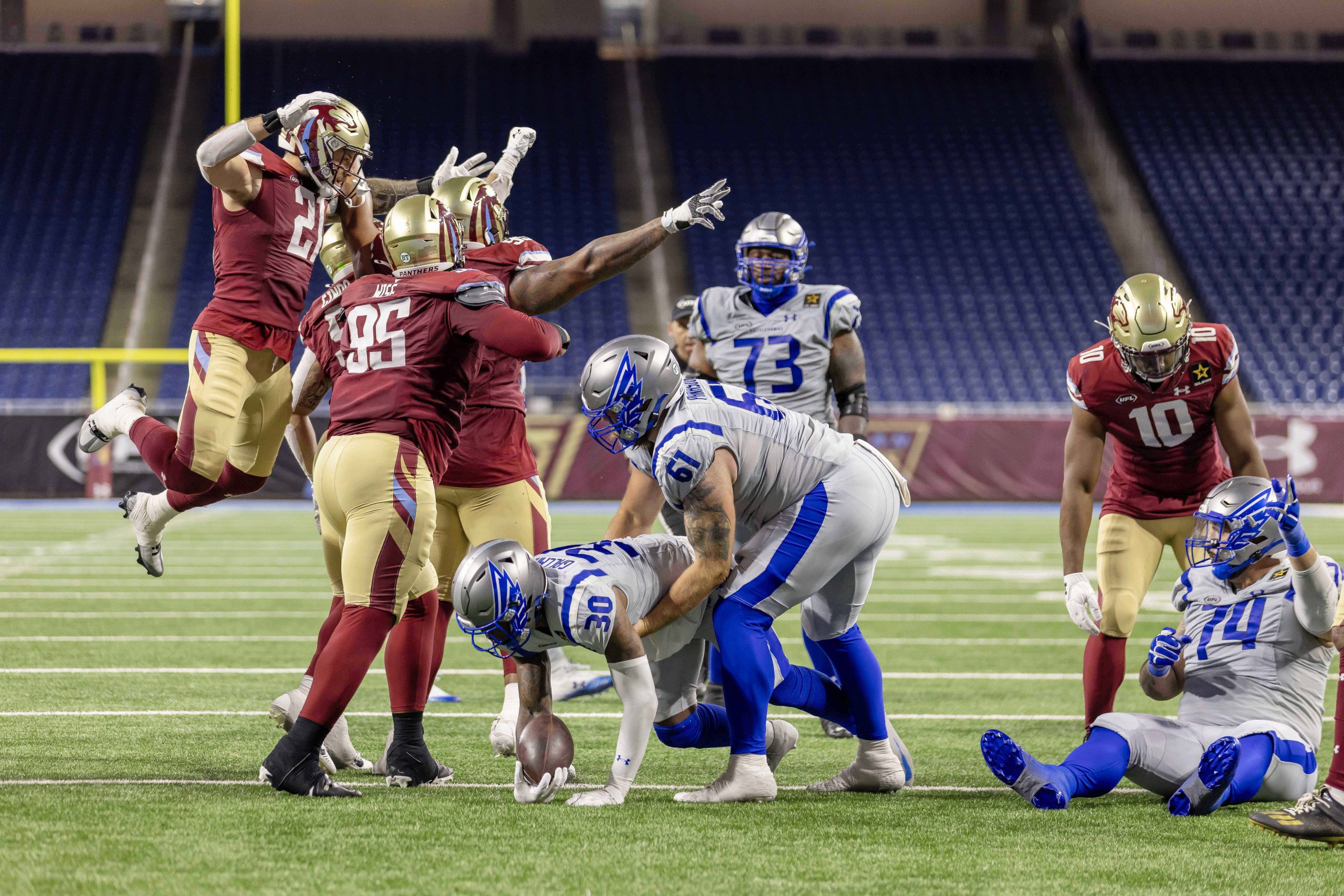 Ein Footballspieler in Blau und Weiß wird angegriffen und verliert den Ball, während vier Spieler in Rot und Gold verteidigen. Ein rot gekleideter Spieler springt mit erhobenen Armen, ein anderer greift nach dem Ball – Nacua schlägt Fan und sorgt für Skandal in den leeren Stadionsitzen. Diese Beschreibung wurde mit der FootballR KI automatisch generiert.