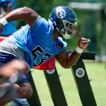Ein Footballspieler, Saahdiq Charles, in blauem Trikot und Helm mit der Nummer 55, übt eine Übung. Er duckt sich tief und schlängelt sich an Tackling-Dummies auf dem Rasen vorbei. Sein konzentrierter Gesichtsausdruck harmoniert perfekt mit den Trainingsgeräten und dem Grün im Hintergrund und vermittelt den Eindruck einer Outdoor-Einheit. Diese Beschreibung wurde mit der FootballR KI automatisch generiert.