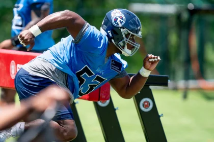 Ein Footballspieler, Saahdiq Charles, in blauem Trikot und Helm mit der Nummer 55, übt eine Übung. Er duckt sich tief und schlängelt sich an Tackling-Dummies auf dem Rasen vorbei. Sein konzentrierter Gesichtsausdruck harmoniert perfekt mit den Trainingsgeräten und dem Grün im Hintergrund und vermittelt den Eindruck einer Outdoor-Einheit. Diese Beschreibung wurde mit der FootballR KI automatisch generiert.
