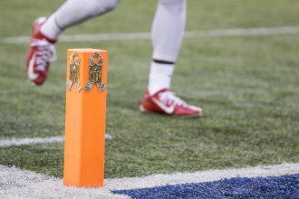 EPA, CPOE, DVOA und Success Rate -Nahaufnahme eines orangefarbenen Football-Pylons mit NFL-Logos und Tarnbändern, der die Seitenlinien während der 18 Spiele NFL Saison markiert. Ein Spieler in roten Stollenschuhen und weißen Hosen sprintet über das Kunstrasenfeld, wo eine weiße Seitenlinie auf eine leuchtend blaue Endzone trifft. Diese Beschreibung wurde mit der FootballR KI automatisch generiert.