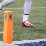 EPA, CPOE, DVOA und Success Rate -Nahaufnahme eines orangefarbenen Football-Pylons mit NFL-Logos und Tarnbändern, der die Seitenlinien während der 18 Spiele NFL Saison markiert. Ein Spieler in roten Stollenschuhen und weißen Hosen sprintet über das Kunstrasenfeld, wo eine weiße Seitenlinie auf eine leuchtend blaue Endzone trifft. Diese Beschreibung wurde mit der FootballR KI automatisch generiert.