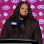 Ashton Gillotte, ein Mann mit langen Locken, steht selbstbewusst in kastanienbraunem Anzug und schwarzem Hemd am Podium. Auf dem kastanienbraunen Hintergrund sind die Logos der Kardinäle und der ACC zu sehen. Vor ihm prangt ein Mikrofon mit dem ACC-Logo. Diese Beschreibung wurde mit der FootballR KI automatisch generiert.