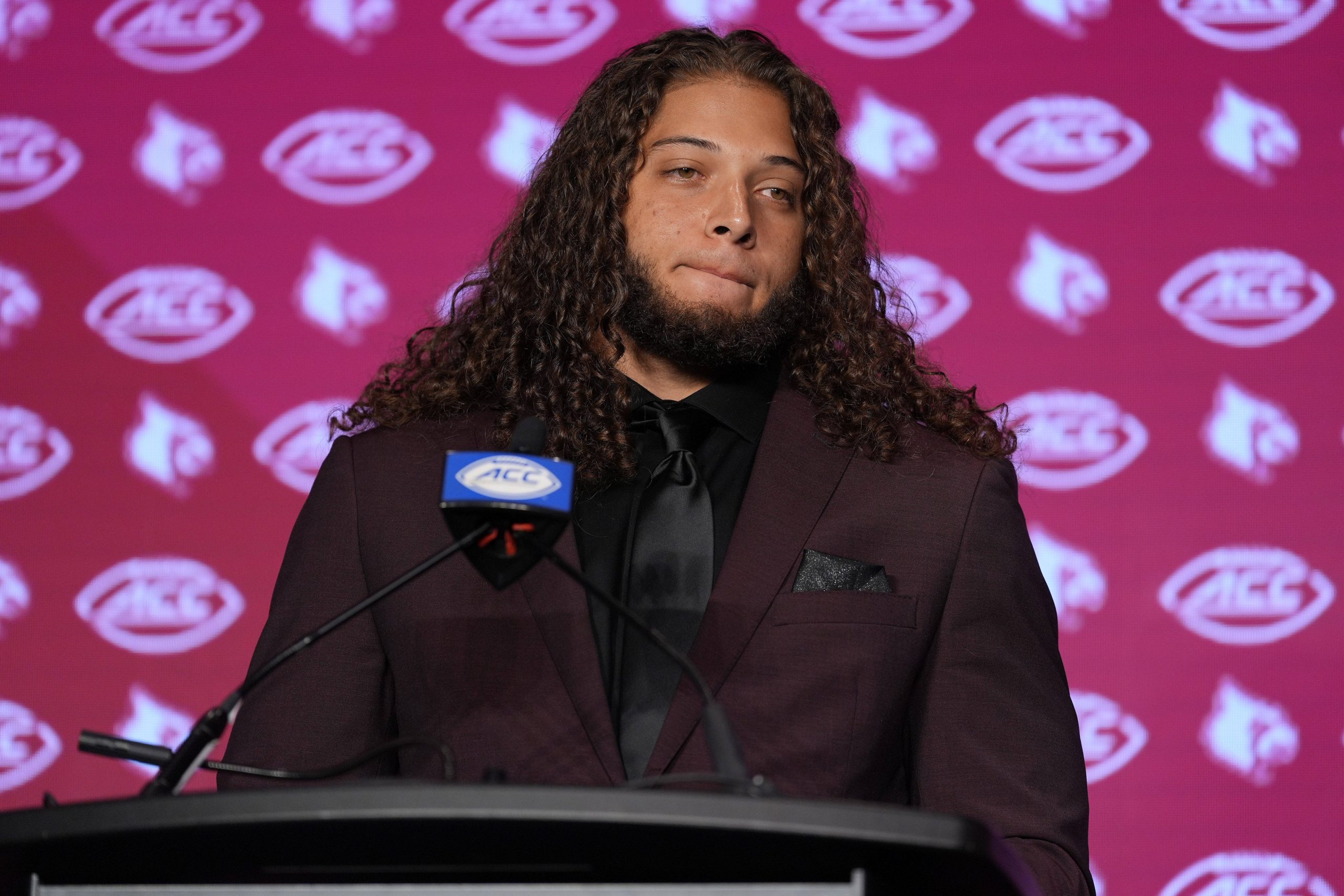 Ashton Gillotte, ein Mann mit langen Locken, steht selbstbewusst in kastanienbraunem Anzug und schwarzem Hemd am Podium. Auf dem kastanienbraunen Hintergrund sind die Logos der Kardinäle und der ACC zu sehen. Vor ihm prangt ein Mikrofon mit dem ACC-Logo. Diese Beschreibung wurde mit der FootballR KI automatisch generiert.