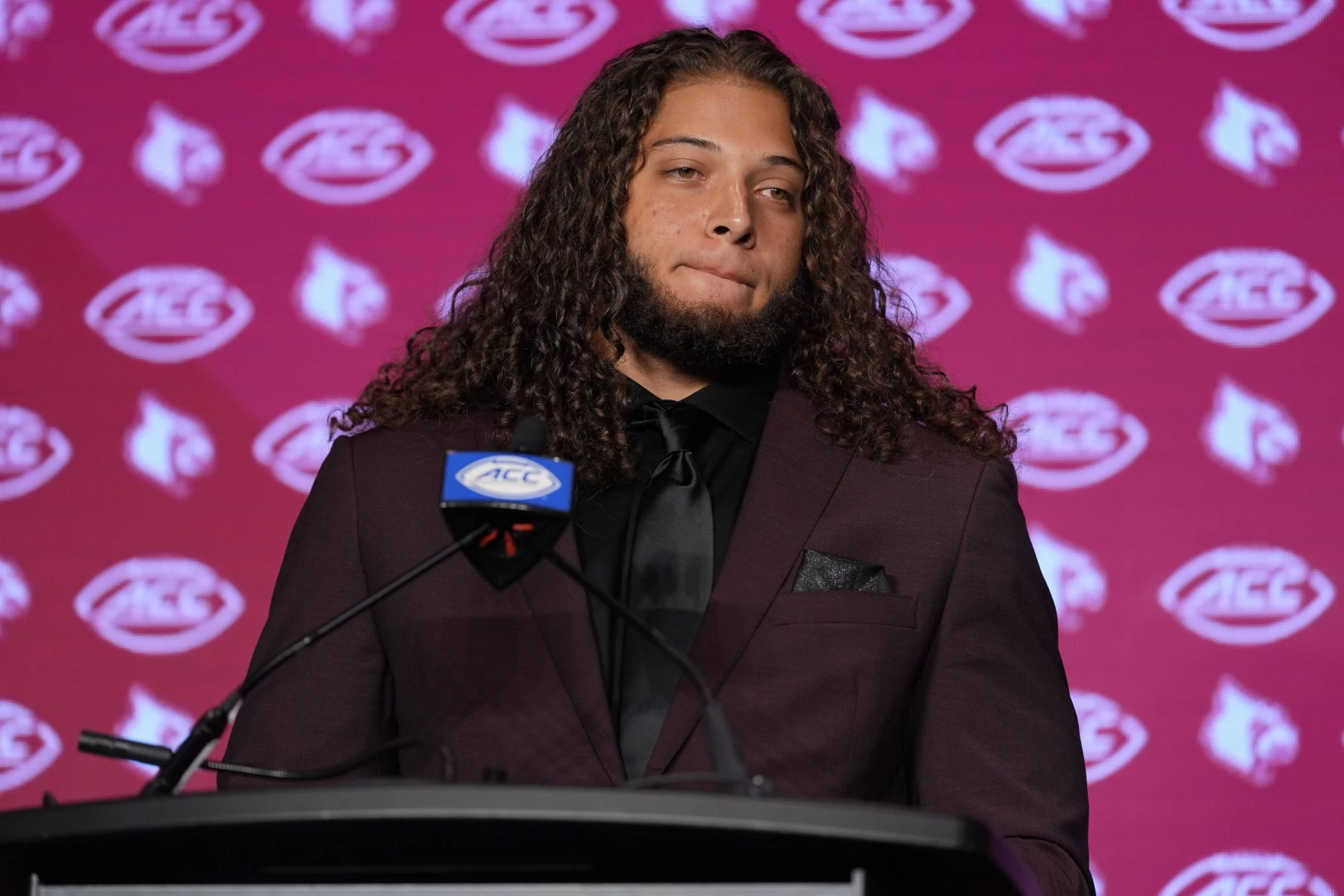 Ashton Gillotte, ein Mann mit langen Locken, steht selbstbewusst in kastanienbraunem Anzug und schwarzem Hemd am Podium. Auf dem kastanienbraunen Hintergrund sind die Logos der Kardinäle und der ACC zu sehen. Vor ihm prangt ein Mikrofon mit dem ACC-Logo. Diese Beschreibung wurde mit der FootballR KI automatisch generiert.