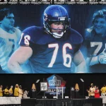 Auf einer großen Leinwand sind Bilder eines American-Football-Spielers im blauen Trikot mit der Nummer 76 zu sehen, der Steve McMichael würdigt. Darunter stehen Menschen in gelben Jacken und beigen Hüten auf der Bühne in der Nähe eines Podiums mit dem Logo der Pro Football Hall of Fame. Diese Beschreibung wurde mit der FootballR KI automatisch generiert.