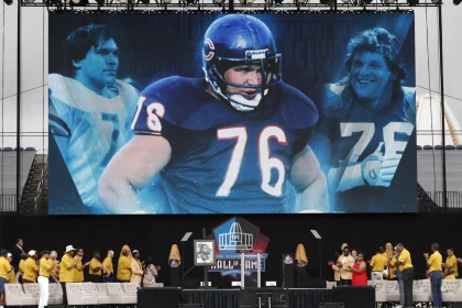 Auf einer großen Leinwand sind Bilder eines American-Football-Spielers im blauen Trikot mit der Nummer 76 zu sehen, der Steve McMichael würdigt. Darunter stehen Menschen in gelben Jacken und beigen Hüten auf der Bühne in der Nähe eines Podiums mit dem Logo der Pro Football Hall of Fame. Diese Beschreibung wurde mit der FootballR KI automatisch generiert.