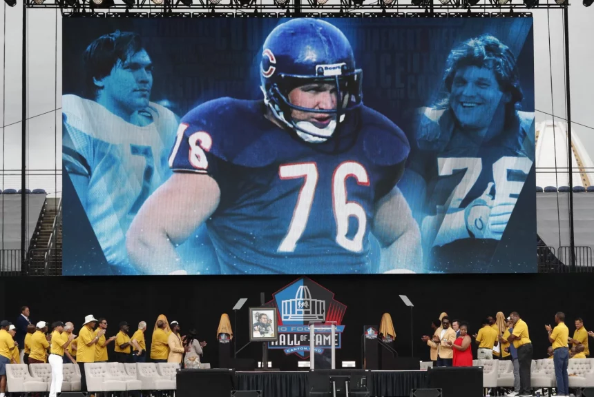 Auf einer großen Leinwand sind Bilder eines American-Football-Spielers im blauen Trikot mit der Nummer 76 zu sehen, der Steve McMichael würdigt. Darunter stehen Menschen in gelben Jacken und beigen Hüten auf der Bühne in der Nähe eines Podiums mit dem Logo der Pro Football Hall of Fame. Diese Beschreibung wurde mit der FootballR KI automatisch generiert.