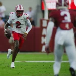 TJ Finley in weiß-roter Uniform (Nummer 2) hält den Ball und rennt über eine Wiese, während TJ Finley (Kastanienbraun-Weiß, Nummer 3) näher kommt. Im Hintergrund sind Zuschauer und Beschilderung unscharf zu sehen. Diese Beschreibung wurde mit der FootballR KI automatisch generiert.