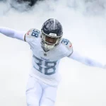 Ein Titans Star betritt das Spielfeld mit ausgestreckten Armen durch eine Rauchwolke. In seinem weißen Trikot mit der Nummer 38, weißen Hosen und einem marineblauen Helm verkörpert er die dynamische und energiegeladene Atmosphäre, die die Essenz des Tennessee Titans Footballs einfängt. Diese Beschreibung wurde mit der FootballR KI automatisch generiert.