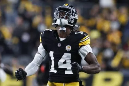 Ein Footballspieler, George Pickens, trägt eine schwarz-gelbe Uniform der Pittsburgh Steelers und den Helm mit der Nummer 14. Er ballt die Fäuste und blickt emotional nach oben auf das Spielfeld. Der unscharfe Hintergrund zeigt ein Stadion mit Fans und Lichtern. Diese Beschreibung wurde mit der FootballR KI automatisch generiert.