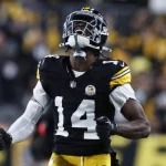 Ein Footballspieler, George Pickens, trägt eine schwarz-gelbe Uniform der Pittsburgh Steelers und den Helm mit der Nummer 14. Er ballt die Fäuste und blickt emotional nach oben auf das Spielfeld. Der unscharfe Hintergrund zeigt ein Stadion mit Fans und Lichtern. Diese Beschreibung wurde mit der FootballR KI automatisch generiert.