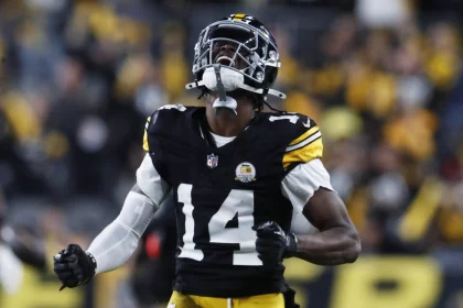 Ein Footballspieler, George Pickens, trägt eine schwarz-gelbe Uniform der Pittsburgh Steelers und den Helm mit der Nummer 14. Er ballt die Fäuste und blickt emotional nach oben auf das Spielfeld. Der unscharfe Hintergrund zeigt ein Stadion mit Fans und Lichtern. Diese Beschreibung wurde mit der FootballR KI automatisch generiert.