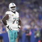 Left Tackle Terron Armstead in weißer Miami-Dolphin-Uniform mit türkisfarbener Hose und Helm steht selbstbewusst auf dem Spielfeld, die Hände in die Hüften gestemmt. Stolz prangt die Nummer 72 auf seinem Trikot, während im verschwommenen Hintergrund des Stadions die Menge jubelt. Diese Beschreibung wurde mit der FootballR KI automatisch generiert.