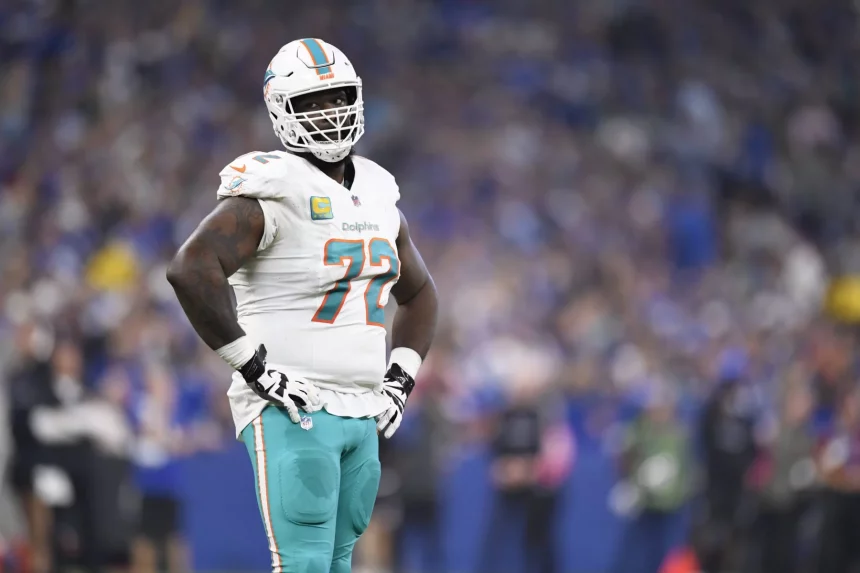 Left Tackle Terron Armstead in weißer Miami-Dolphin-Uniform mit türkisfarbener Hose und Helm steht selbstbewusst auf dem Spielfeld, die Hände in die Hüften gestemmt. Stolz prangt die Nummer 72 auf seinem Trikot, während im verschwommenen Hintergrund des Stadions die Menge jubelt. Diese Beschreibung wurde mit der FootballR KI automatisch generiert.