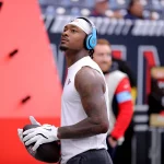 Diggs Deal: Stefon Diggs in ärmellosem weißen Trikot und Stirnband steht da und hält einen Football mit der Aufschrift „Diggs-Deal“. Er trägt blaue Kopfhörer, auf seinen Armen sind Tattoos zu sehen. Die verschwommenen Zuschauer und Strukturen im Stadion lassen die Stimmung vor dem Spiel oder Training erahnen und unterstreichen die Konzentration des Sportlers. Diese Beschreibung wurde mit der FootballR KI automatisch generiert.