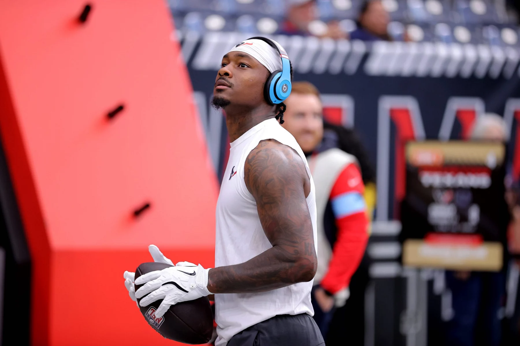 Diggs Deal: Stefon Diggs in ärmellosem weißen Trikot und Stirnband steht da und hält einen Football mit der Aufschrift „Diggs-Deal“. Er trägt blaue Kopfhörer, auf seinen Armen sind Tattoos zu sehen. Die verschwommenen Zuschauer und Strukturen im Stadion lassen die Stimmung vor dem Spiel oder Training erahnen und unterstreichen die Konzentration des Sportlers. Diese Beschreibung wurde mit der FootballR KI automatisch generiert.
