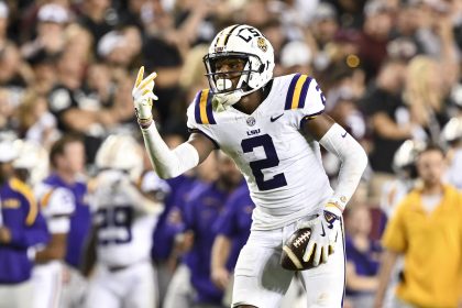 Ein Footballspieler, Kyren Lacy, in weißer Uniform mit violetten und gelben Akzenten und einem Helm mit der Aufschrift „LSU“, hält einen Football. Mit der linken Hand streckt er zwei Finger nach oben. Im Hintergrund sind Zuschauer zu sehen, einige in leuchtend gelber und violetter Kleidung. Diese Beschreibung wurde mit der FootballR KI automatisch generiert.