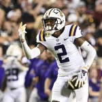 Ein Footballspieler, Kyren Lacy, in weißer Uniform mit violetten und gelben Akzenten und einem Helm mit der Aufschrift „LSU“, hält einen Football. Mit der linken Hand streckt er zwei Finger nach oben. Im Hintergrund sind Zuschauer zu sehen, einige in leuchtend gelber und violetter Kleidung. Diese Beschreibung wurde mit der FootballR KI automatisch generiert.