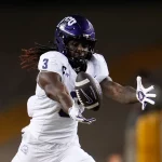 TCU Wide Receiver Savion Williams, in weißer Uniform und lila Helm mit der Nummer 3, streckt während eines Spiels die Arme aus, um einen Football in der Luft zu fangen. Sein Blick ist auf den Ball gerichtet, während die Stadionlichter die Szene erhellen. Diese Beschreibung wurde mit der FootballR KI automatisch generiert.