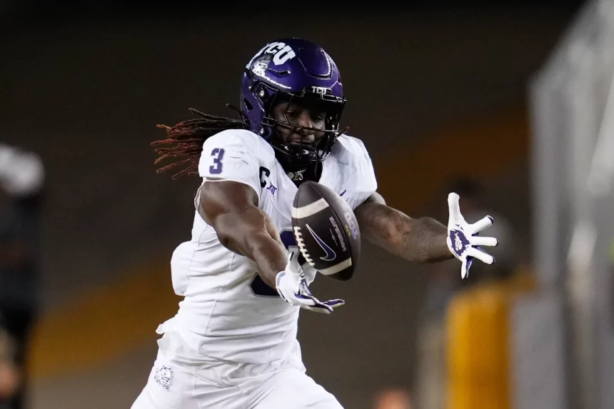 TCU Wide Receiver Savion Williams, in weißer Uniform und lila Helm mit der Nummer 3, streckt während eines Spiels die Arme aus, um einen Football in der Luft zu fangen. Sein Blick ist auf den Ball gerichtet, während die Stadionlichter die Szene erhellen. Diese Beschreibung wurde mit der FootballR KI automatisch generiert.