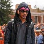 Ein Mann in schwarzer Kapuzenjacke, rosa Beanie und orangefarbenen Kopfhörern läuft draußen inmitten einer Menschenmenge. Er trägt eine Sonnenbrille und einen Rucksack. Hinter Absperrungen stehen Fans in orangefarbener Kleidung – möglicherweise warten sie auf Nico Iamaleava Wechsel. Diese Beschreibung wurde mit der FootballR KI automatisch generiert.