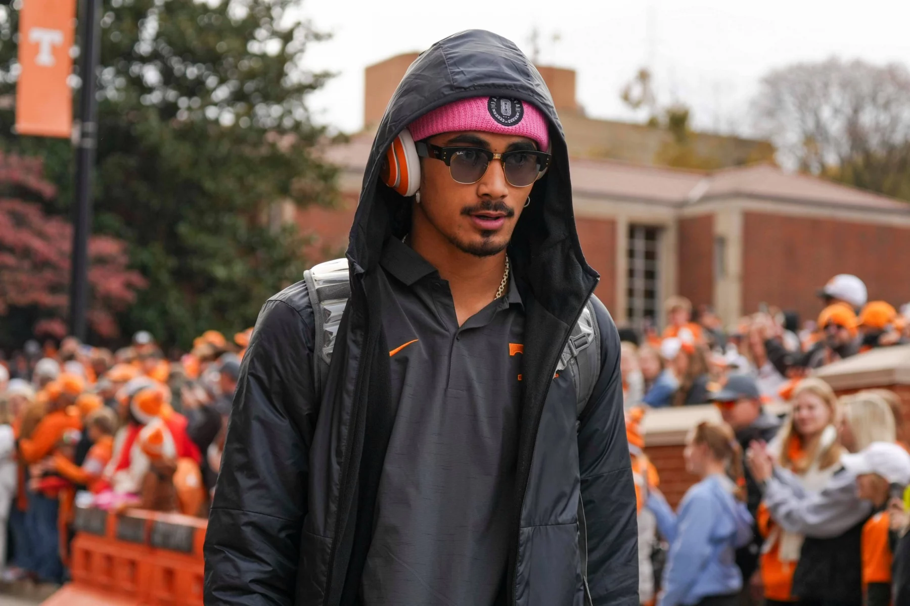 Ein Mann in schwarzer Kapuzenjacke, rosa Beanie und orangefarbenen Kopfhörern läuft draußen inmitten einer Menschenmenge. Er trägt eine Sonnenbrille und einen Rucksack. Hinter Absperrungen stehen Fans in orangefarbener Kleidung – möglicherweise warten sie auf Nico Iamaleava Wechsel. Diese Beschreibung wurde mit der FootballR KI automatisch generiert.