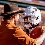 Isaiah Bond - Ein Mann mit braunem Hut und Hemd umarmt Isaiah Bond herzlich. Er trägt eine orange-weiße Uniform und einen Helm mit Longhorn-Logo. Sie stehen auf dem Footballfeld, umgeben von verschwommenen Zuschauern, die die lebhafte Spieltagsatmosphäre einfangen. Diese Beschreibung wurde mit der FootballR KI automatisch generiert.